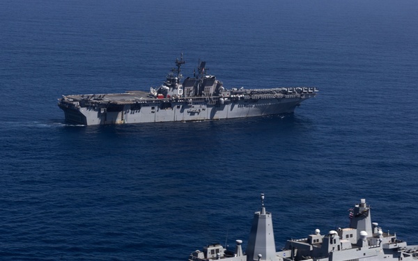Two U.S. Navy warships are underway in open blue ocean waters. The foreground vessel appears to be a San Antonio-class amphibious transport dock (LPD) with helicopters visible on its flight deck, while the background vessel is a larger Wasp-class or America-class amphibious assault ship (LHA/LHD). Both ships are sailing in formation, suggesting a coordinated naval exercise or transit operation.