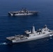 Two U.S. Navy warships are underway in open blue ocean waters. The foreground vessel appears to be a San Antonio-class amphibious transport dock (LPD) with helicopters visible on its flight deck, while the background vessel is a larger Wasp-class or America-class amphibious assault ship (LHA/LHD). Both ships are sailing in formation, suggesting a coordinated naval exercise or transit operation.
