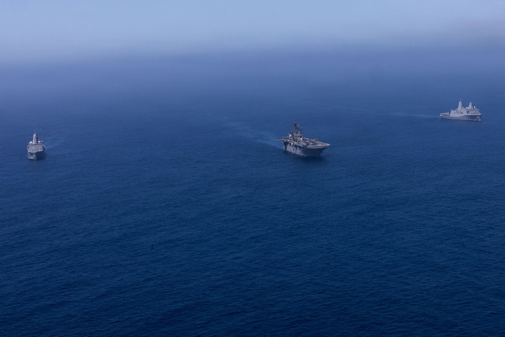 An aerial photograph shows three naval vessels sailing in formation across open deep-blue ocean water. The center vessel appears to be a large amphibious assault ship with a flat deck, flanked by two smaller warships on the left and right. The image is taken from a high altitude with hazy atmospheric conditions reducing visibility at the horizon.