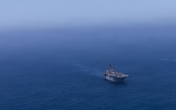 An aerial photograph shows three naval vessels sailing in formation across open deep-blue ocean water. The center vessel appears to be a large amphibious assault ship with a flat deck, flanked by two smaller warships on the left and right. The image is taken from a high altitude with hazy atmospheric conditions reducing visibility at the horizon.