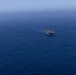 An aerial photograph shows three naval vessels sailing in formation across open deep-blue ocean water. The center vessel appears to be a large amphibious assault ship with a flat deck, flanked by two smaller warships on the left and right. The image is taken from a high altitude with hazy atmospheric conditions reducing visibility at the horizon.