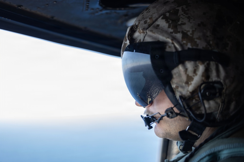 A military personnel member is photographed in close profile view from inside an aircraft, looking out through an open window or door at what appears to be sky and water below. The individual is wearing full flight/combat gear including a camouflage helmet, tinted visor, oxygen or communications mask, and hearing protection. The image captures a moment of aerial observation during what appears to be a military flight operation.