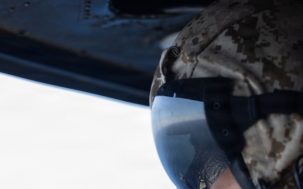 A military personnel member is photographed in close profile view from inside an aircraft, looking out through an open window or door at what appears to be sky and water below. The individual is wearing full flight/combat gear including a camouflage helmet, tinted visor, oxygen or communications mask, and hearing protection. The image captures a moment of aerial observation during what appears to be a military flight operation.