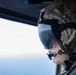 A military personnel member is photographed in close profile view from inside an aircraft, looking out through an open window or door at what appears to be sky and water below. The individual is wearing full flight/combat gear including a camouflage helmet, tinted visor, oxygen or communications mask, and hearing protection. The image captures a moment of aerial observation during what appears to be a military flight operation.