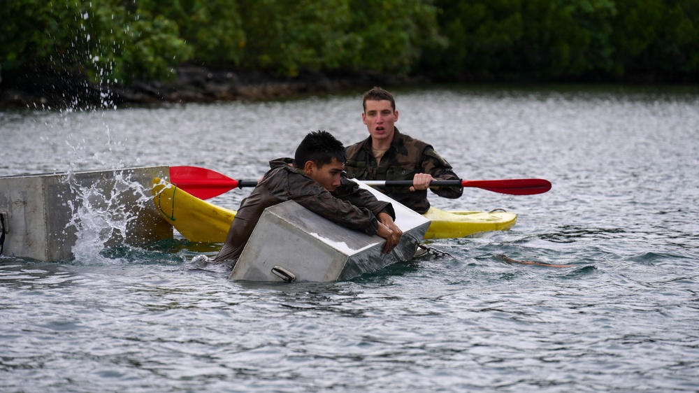 Two individuals in military-style camouflage clothing are engaged in a water-based training exercise. One person is clinging to a partially submerged metal container or box in the water, while a second person paddles a yellow kayak with a red paddle nearby. The scene appears to be a military or survival training drill taking place on a calm body of water with dense green vegetation in the background.