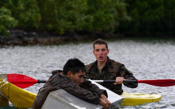 Two individuals in military-style camouflage clothing are engaged in a water-based training exercise. One person is clinging to a partially submerged metal container or box in the water, while a second person paddles a yellow kayak with a red paddle nearby. The scene appears to be a military or survival training drill taking place on a calm body of water with dense green vegetation in the background.