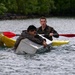 Two individuals in military-style camouflage clothing are engaged in a water-based training exercise. One person is clinging to a partially submerged metal container or box in the water, while a second person paddles a yellow kayak with a red paddle nearby. The scene appears to be a military or survival training drill taking place on a calm body of water with dense green vegetation in the background.