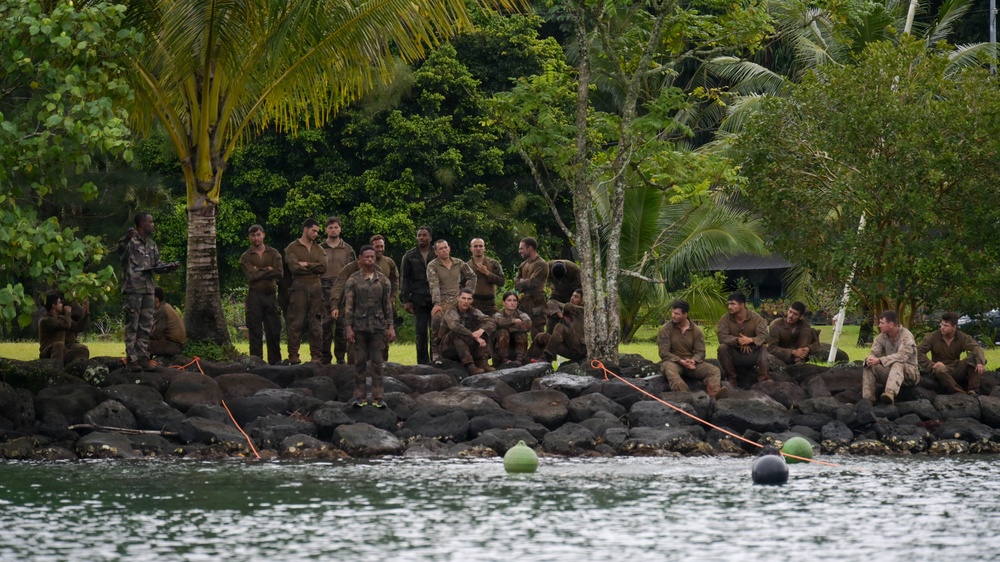 A large group of approximately 20 individuals wearing matching olive/brown uniforms are gathered on a rocky shoreline bordered by tropical vegetation. They appear to be resting or receiving instruction near a body of water, with orange ropes and buoys visible in the foreground. The tropical setting with palm trees and dense greenery suggests a warm, island-like environment.