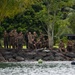 A large group of approximately 20 individuals wearing matching olive/brown uniforms are gathered on a rocky shoreline bordered by tropical vegetation. They appear to be resting or receiving instruction near a body of water, with orange ropes and buoys visible in the foreground. The tropical setting with palm trees and dense greenery suggests a warm, island-like environment.