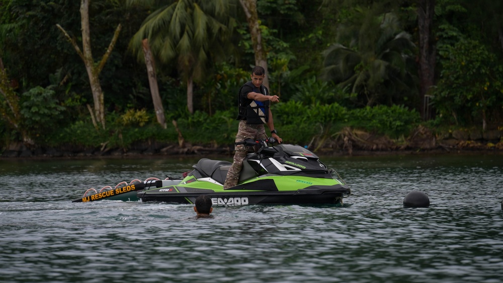 A person in military-style camouflage pants and a black shirt stands on a green and black Sea-Doo personal watercraft on a calm body of water. The jet ski is equipped with a rescue sled labeled 'MJ Rescue Sleds,' indicating a water rescue training or operational context. A swimmer's head is visible in the water nearby, suggesting an active rescue drill or demonstration.