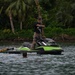A person in military-style camouflage pants and a black shirt stands on a green and black Sea-Doo personal watercraft on a calm body of water. The jet ski is equipped with a rescue sled labeled 'MJ Rescue Sleds,' indicating a water rescue training or operational context. A swimmer's head is visible in the water nearby, suggesting an active rescue drill or demonstration.