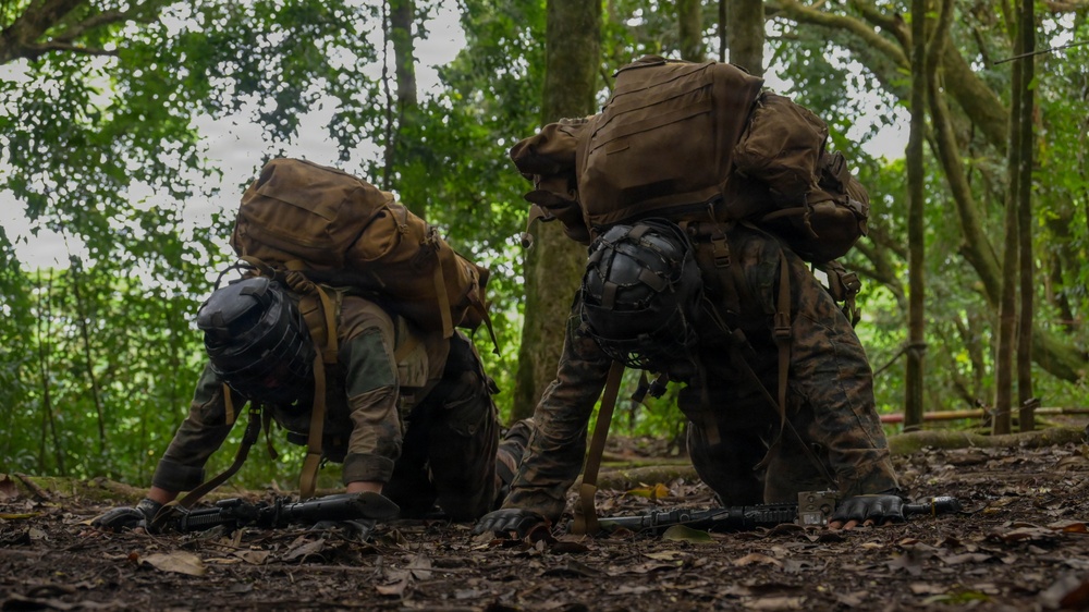 Two military personnel in full combat gear are crawling or low-crawling through a forested jungle environment. Both are equipped with large tactical backpacks, helmets, gloves, and rifles visible on the ground beneath them. The scene appears to depict a military training exercise or field operation in a dense tropical woodland setting.