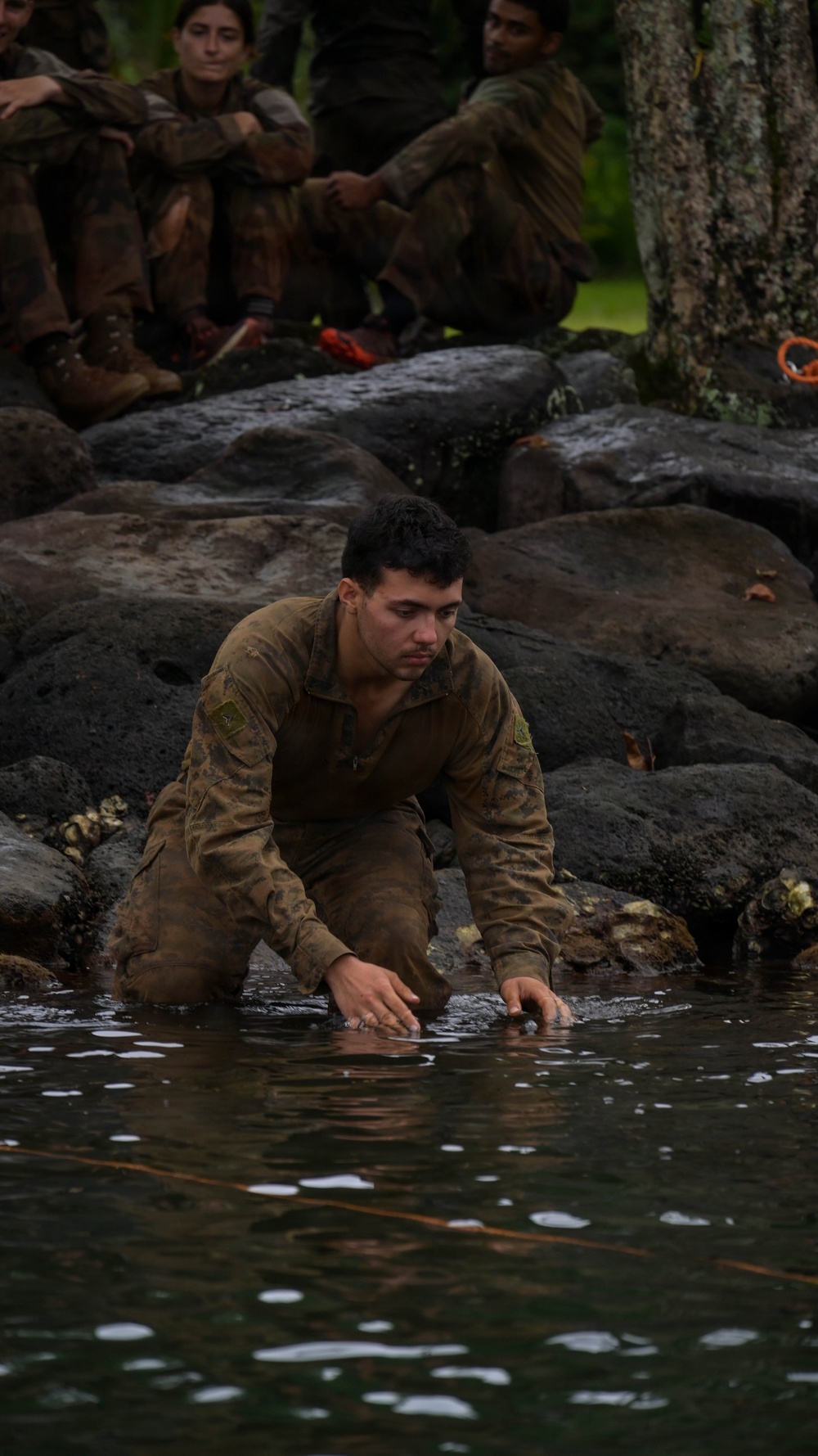 A soldier in olive/brown military fatigues is kneeling at the edge of a dark water body, with hands touching or reaching into the water. Several other soldiers in similar uniforms are seated on dark volcanic rocks in the background, observing. The setting appears to be a jungle or tropical environment, suggesting a military training exercise.