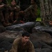 A soldier in olive/brown military fatigues is kneeling at the edge of a dark water body, with hands touching or reaching into the water. Several other soldiers in similar uniforms are seated on dark volcanic rocks in the background, observing. The setting appears to be a jungle or tropical environment, suggesting a military training exercise.