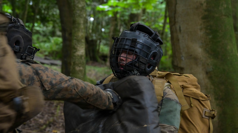 Two individuals in military camouflage uniforms are engaged in a close-quarters combat training exercise in a forested environment. One person is wearing a black protective headgear with a metal cage face guard, indicating a controlled sparring or martial arts drill. The setting and equipment suggest this is an organized military hand-to-hand combat training session.