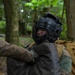 Two individuals in military camouflage uniforms are engaged in a close-quarters combat training exercise in a forested environment. One person is wearing a black protective headgear with a metal cage face guard, indicating a controlled sparring or martial arts drill. The setting and equipment suggest this is an organized military hand-to-hand combat training session.