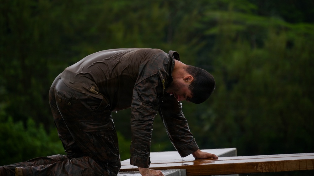 A man is working outdoors in heavy rain, leaning over what appears to be a wooden structure or surface. He is thoroughly soaked, wearing a dark wax-style jacket that is visibly wet and glistening. The background features dense green forest trees, suggesting a rural or woodland setting.