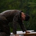 A man is working outdoors in heavy rain, leaning over what appears to be a wooden structure or surface. He is thoroughly soaked, wearing a dark wax-style jacket that is visibly wet and glistening. The background features dense green forest trees, suggesting a rural or woodland setting.
