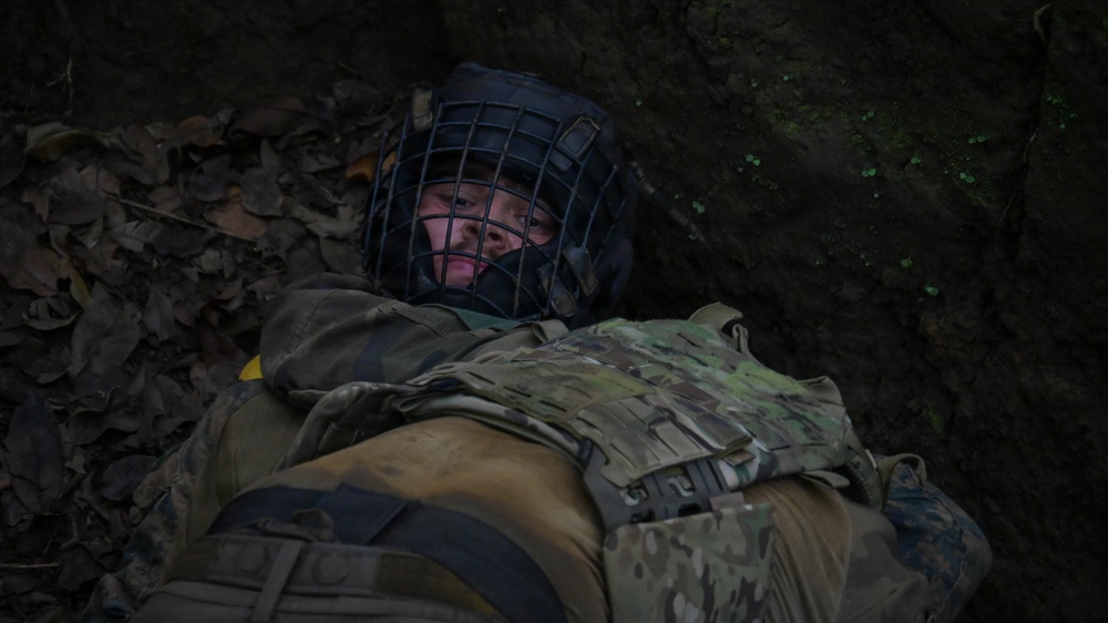 A person wearing military tactical gear and a protective face cage helmet is lying on the ground among dead leaves, appearing to be in a low crawl or prone position near a dirt embankment. The individual is equipped with a multicam-pattern plate carrier vest and dark outer clothing. The setting appears to be an outdoor woodland or forested training environment.