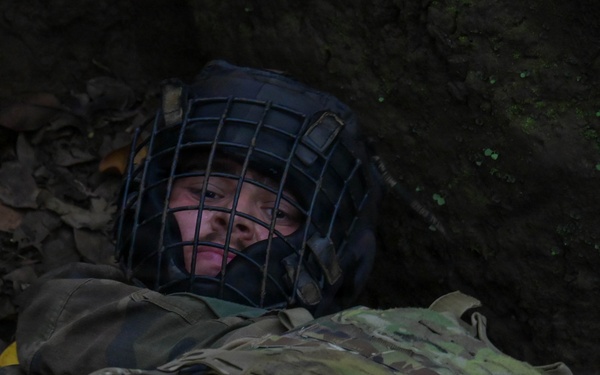 A person wearing military tactical gear and a protective face cage helmet is lying on the ground among dead leaves, appearing to be in a low crawl or prone position near a dirt embankment. The individual is equipped with a multicam-pattern plate carrier vest and dark outer clothing. The setting appears to be an outdoor woodland or forested training environment.