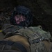 A person wearing military tactical gear and a protective face cage helmet is lying on the ground among dead leaves, appearing to be in a low crawl or prone position near a dirt embankment. The individual is equipped with a multicam-pattern plate carrier vest and dark outer clothing. The setting appears to be an outdoor woodland or forested training environment.