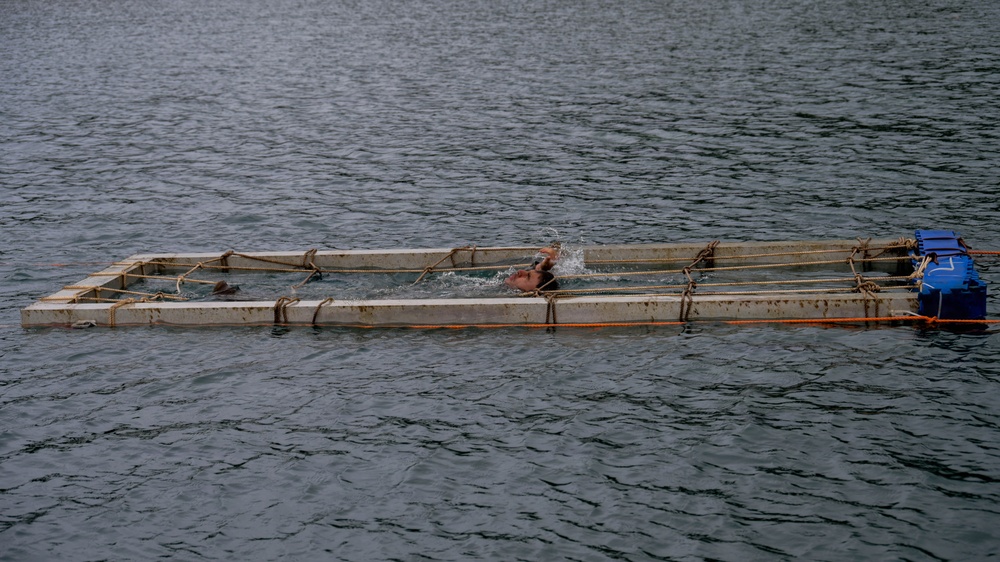 A person is swimming inside a rectangular floating frame structure on open water. The structure appears to be a makeshift floating pen or enclosure constructed from concrete or metal beams, secured with ropes and orange cordage. A blue container or equipment box is attached to the right side of the structure.