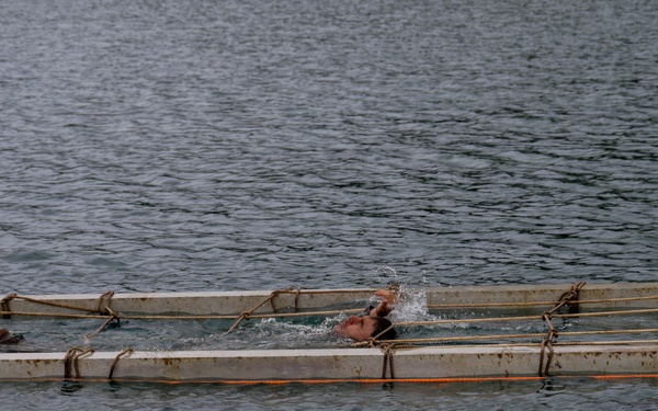 A person is swimming inside a rectangular floating frame structure on open water. The structure appears to be a makeshift floating pen or enclosure constructed from concrete or metal beams, secured with ropes and orange cordage. A blue container or equipment box is attached to the right side of the structure.
