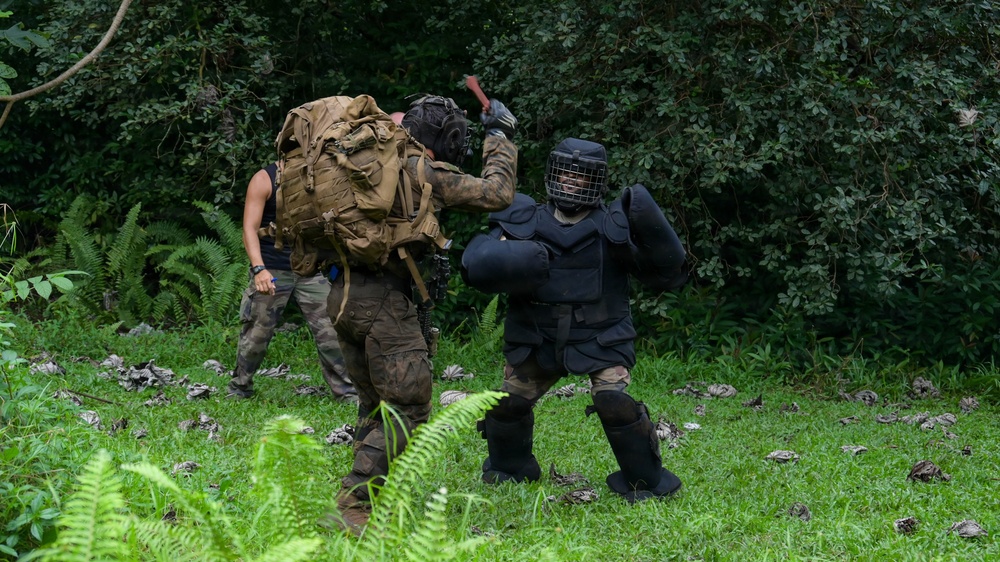 The image shows what appears to be a military or law enforcement training exercise taking place in a grassy outdoor area with dense forest in the background. One person wearing full protective padding and a face cage (a 'red man' or FIST suit) is engaged in a simulated combat scenario with a person in camouflage gear carrying a large tactical backpack. A third individual in civilian-style clothing observes or participates in the background.