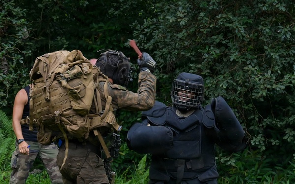 The image shows what appears to be a military or law enforcement training exercise taking place in a grassy outdoor area with dense forest in the background. One person wearing full protective padding and a face cage (a 'red man' or FIST suit) is engaged in a simulated combat scenario with a person in camouflage gear carrying a large tactical backpack. A third individual in civilian-style clothing observes or participates in the background.