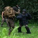 The image shows what appears to be a military or law enforcement training exercise taking place in a grassy outdoor area with dense forest in the background. One person wearing full protective padding and a face cage (a 'red man' or FIST suit) is engaged in a simulated combat scenario with a person in camouflage gear carrying a large tactical backpack. A third individual in civilian-style clothing observes or participates in the background.