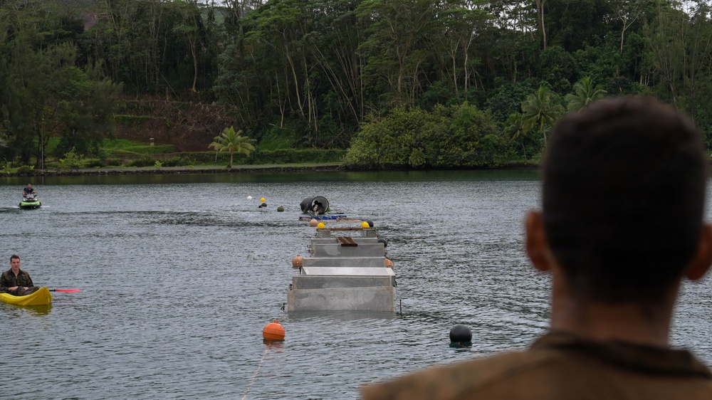 The image shows a military or engineering exercise taking place on a calm inland waterway surrounded by dense tropical vegetation. A floating pontoon bridge or modular floating structure extends across the water, with personnel and equipment visible on and around it. Support watercraft including a kayak and a personal watercraft are positioned nearby, while an observer in military-style clothing watches from the foreground.