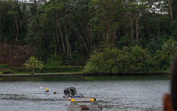 The image shows a military or engineering exercise taking place on a calm inland waterway surrounded by dense tropical vegetation. A floating pontoon bridge or modular floating structure extends across the water, with personnel and equipment visible on and around it. Support watercraft including a kayak and a personal watercraft are positioned nearby, while an observer in military-style clothing watches from the foreground.