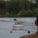 The image shows a military or engineering exercise taking place on a calm inland waterway surrounded by dense tropical vegetation. A floating pontoon bridge or modular floating structure extends across the water, with personnel and equipment visible on and around it. Support watercraft including a kayak and a personal watercraft are positioned nearby, while an observer in military-style clothing watches from the foreground.