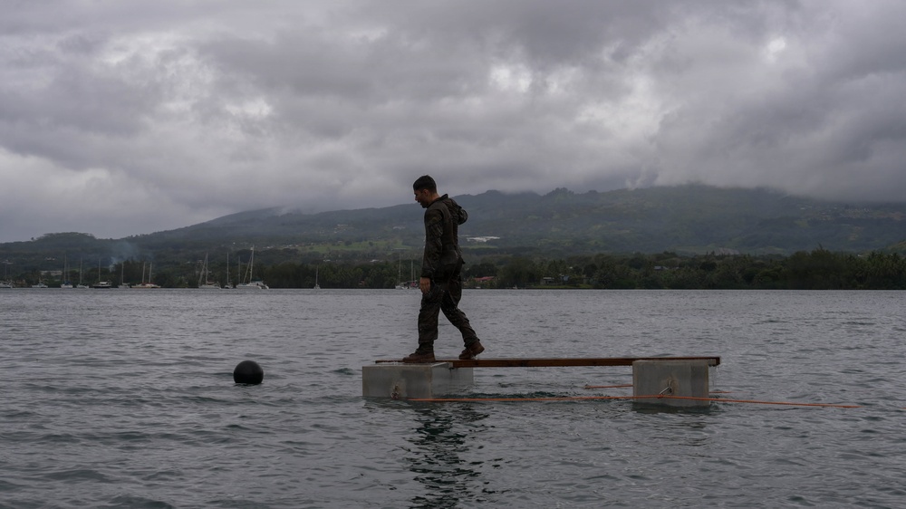 A person dressed in dark military-style clothing stands on a small floating platform in a calm body of water. The scene is set against a backdrop of green, cloud-covered mountains and a gray overcast sky. Several sailboats are visible in the distance near the shoreline.