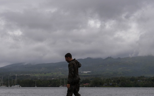 A person dressed in dark military-style clothing stands on a small floating platform in a calm body of water. The scene is set against a backdrop of green, cloud-covered mountains and a gray overcast sky. Several sailboats are visible in the distance near the shoreline.