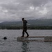 A person dressed in dark military-style clothing stands on a small floating platform in a calm body of water. The scene is set against a backdrop of green, cloud-covered mountains and a gray overcast sky. Several sailboats are visible in the distance near the shoreline.
