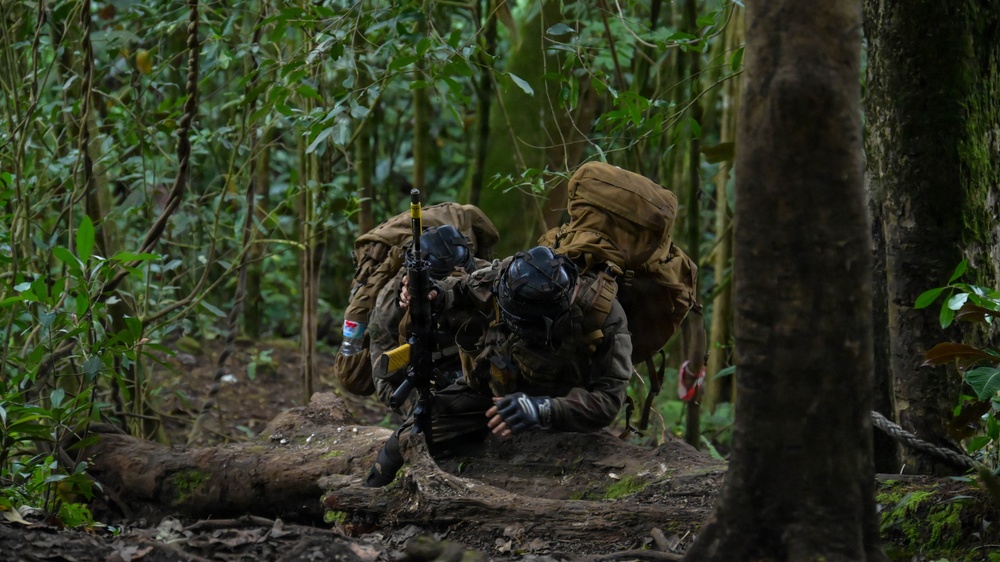 Two heavily equipped individuals in tactical gear and gas masks are crouched behind a fallen log in a dense tropical jungle environment. They appear to be in a defensive or concealed position, with at least one individual holding a firearm. The setting and equipment suggest a military training exercise or tactical operation in a jungle terrain.