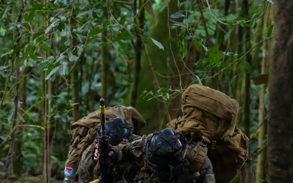Two heavily equipped individuals in tactical gear and gas masks are crouched behind a fallen log in a dense tropical jungle environment. They appear to be in a defensive or concealed position, with at least one individual holding a firearm. The setting and equipment suggest a military training exercise or tactical operation in a jungle terrain.
