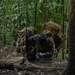 Two heavily equipped individuals in tactical gear and gas masks are crouched behind a fallen log in a dense tropical jungle environment. They appear to be in a defensive or concealed position, with at least one individual holding a firearm. The setting and equipment suggest a military training exercise or tactical operation in a jungle terrain.