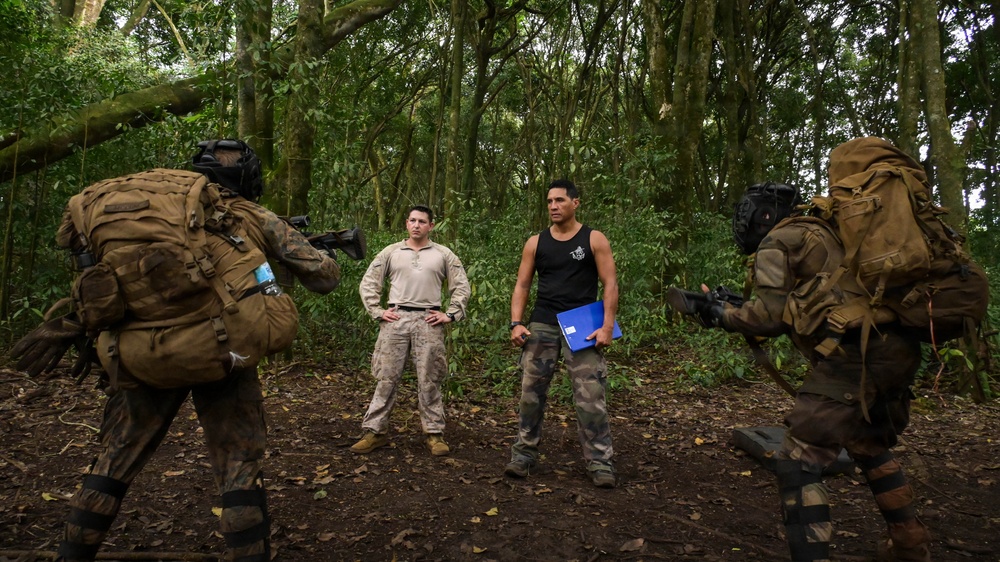 The image shows a jungle or dense forest setting where two heavily equipped soldiers in camouflage gear and full tactical kit are pointing weapons at two individuals standing between them. One of the standing individuals wears a tan military-style shirt with camouflage pants, while the other wears a black sleeveless shirt with camouflage pants and holds a blue folder. The scene appears to depict a military training exercise or scenario.