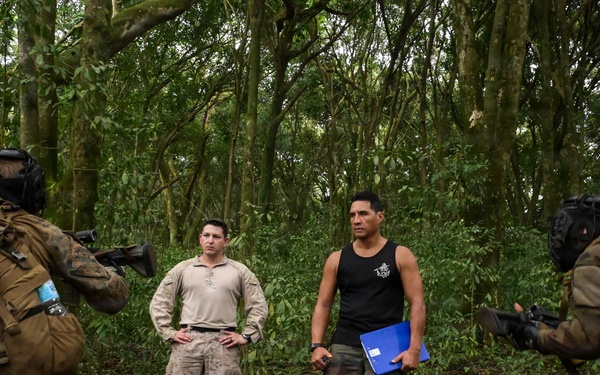 The image shows a jungle or dense forest setting where two heavily equipped soldiers in camouflage gear and full tactical kit are pointing weapons at two individuals standing between them. One of the standing individuals wears a tan military-style shirt with camouflage pants, while the other wears a black sleeveless shirt with camouflage pants and holds a blue folder. The scene appears to depict a military training exercise or scenario.