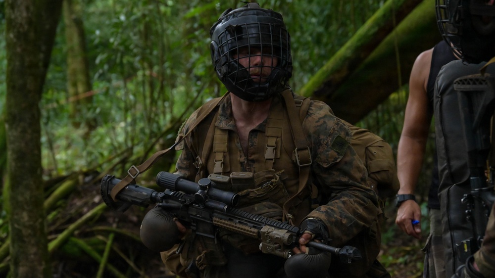 A soldier in woodland camouflage uniform is positioned in a dense jungle environment, holding a scoped assault rifle. They are wearing a black protective face cage/mask over their head, suggesting a training or force-on-force exercise scenario. A second partially visible individual in tactical gear stands to the right.