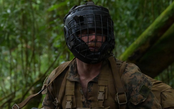 A soldier in woodland camouflage uniform is positioned in a dense jungle environment, holding a scoped assault rifle. They are wearing a black protective face cage/mask over their head, suggesting a training or force-on-force exercise scenario. A second partially visible individual in tactical gear stands to the right.