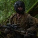 A soldier in woodland camouflage uniform is positioned in a dense jungle environment, holding a scoped assault rifle. They are wearing a black protective face cage/mask over their head, suggesting a training or force-on-force exercise scenario. A second partially visible individual in tactical gear stands to the right.