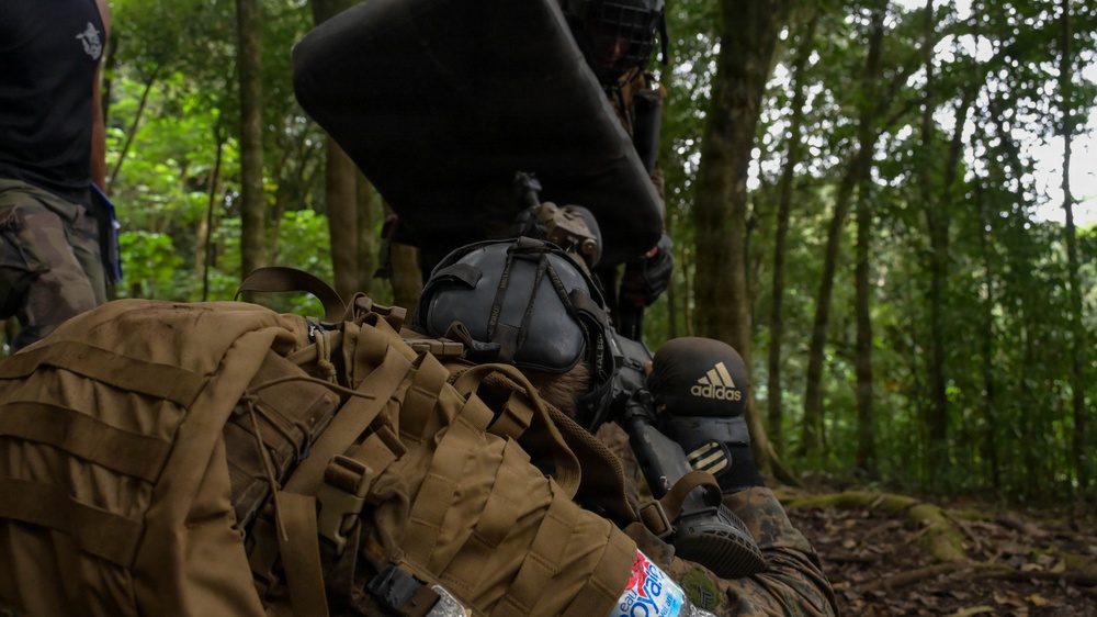 The image shows individuals in a forested environment wearing tactical gear, protective equipment, and carrying backpacks. One person is in a prone position aiming what appears to be an airsoft or paintball rifle, wearing an Adidas-branded protective mask. The setting and equipment suggest this is a recreational airsoft or tactical training exercise rather than a military operation.