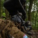 The image shows individuals in a forested environment wearing tactical gear, protective equipment, and carrying backpacks. One person is in a prone position aiming what appears to be an airsoft or paintball rifle, wearing an Adidas-branded protective mask. The setting and equipment suggest this is a recreational airsoft or tactical training exercise rather than a military operation.