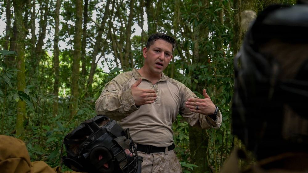 A military personnel member in tan/desert camouflage uniform is speaking and gesturing with both hands in a forested jungle environment. He appears to be briefing or instructing others, as suggested by his animated hand gestures and the presence of at least one other person in the foreground. The setting suggests a field training exercise or operational briefing in a tropical or subtropical woodland area.