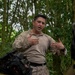 A military personnel member in tan/desert camouflage uniform is speaking and gesturing with both hands in a forested jungle environment. He appears to be briefing or instructing others, as suggested by his animated hand gestures and the presence of at least one other person in the foreground. The setting suggests a field training exercise or operational briefing in a tropical or subtropical woodland area.