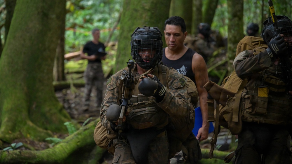 The image depicts a military training exercise in a dense forest environment. Personnel in camouflage uniforms and tactical gear are moving through the wooded terrain, with one individual wearing a protective face cage and boxing gloves, suggesting a combatives or hand-to-hand combat training scenario. Civilian-clothed individuals appear to be acting as role players or observers/instructors.