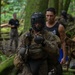 The image depicts a military training exercise in a dense forest environment. Personnel in camouflage uniforms and tactical gear are moving through the wooded terrain, with one individual wearing a protective face cage and boxing gloves, suggesting a combatives or hand-to-hand combat training scenario. Civilian-clothed individuals appear to be acting as role players or observers/instructors.